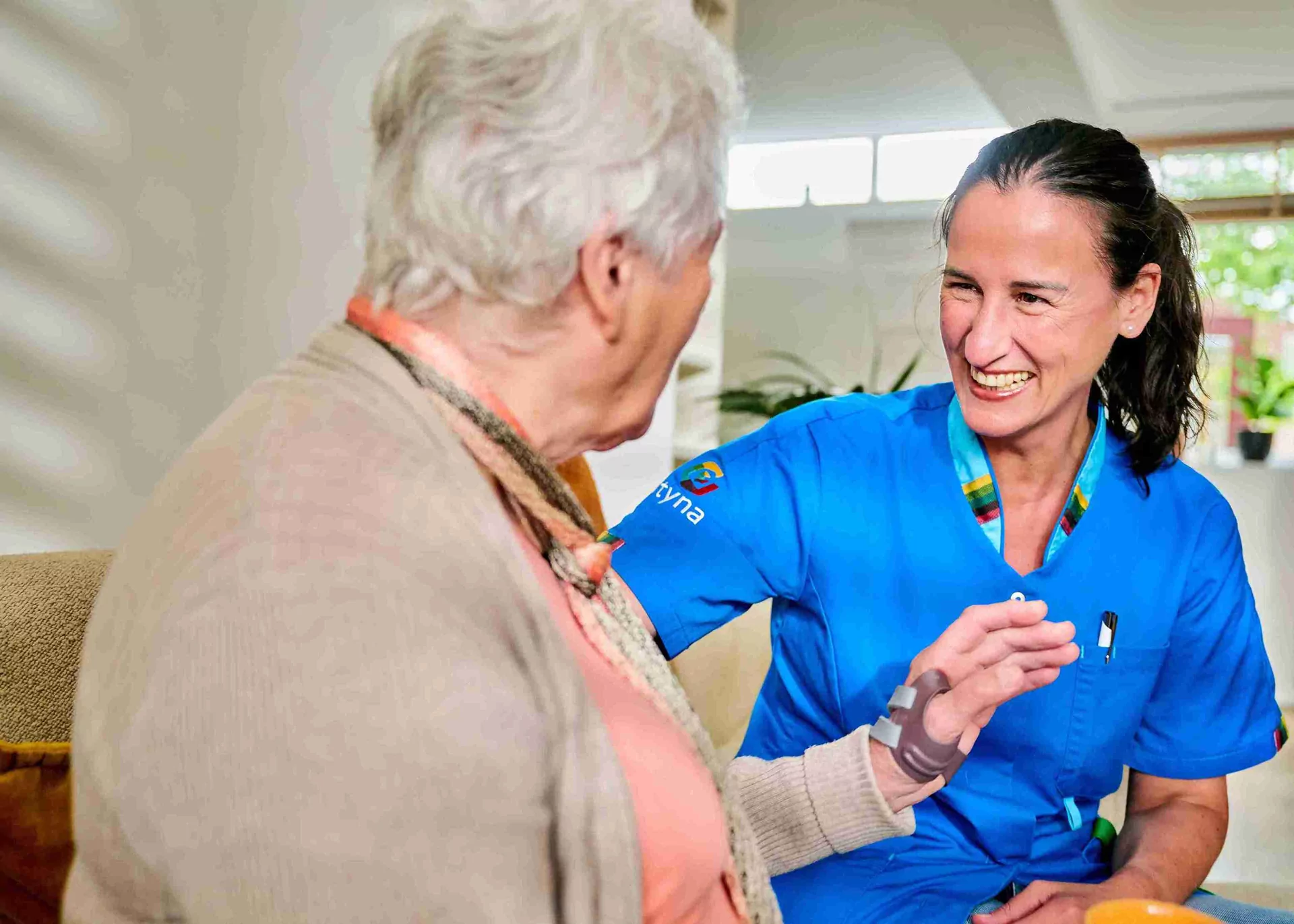 Een verpleegster in blauwe scrubs glimlacht hartelijk terwijl ze in interactie is met een oudere vrouw met grijs haar, die op een bank zit. Ze lijken een vriendelijk en ondersteunend gesprek te hebben in een lichte kamer.