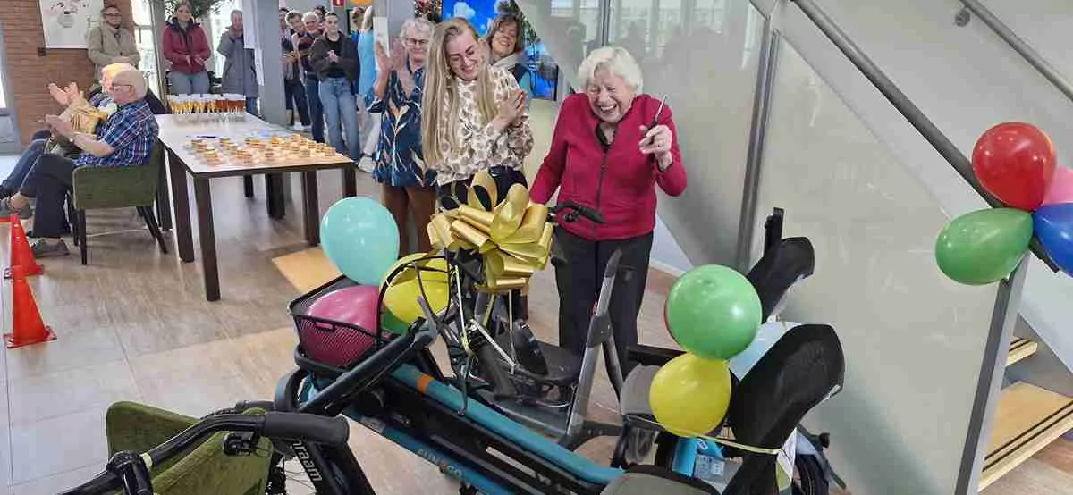 Een oudere vrouw glimlacht en wijst opgewonden naar een versierde driewieler met ballonnen en een gouden strik terwijl mensen om haar heen applaudisseren en feestvieren in een heldere binnenomgeving.