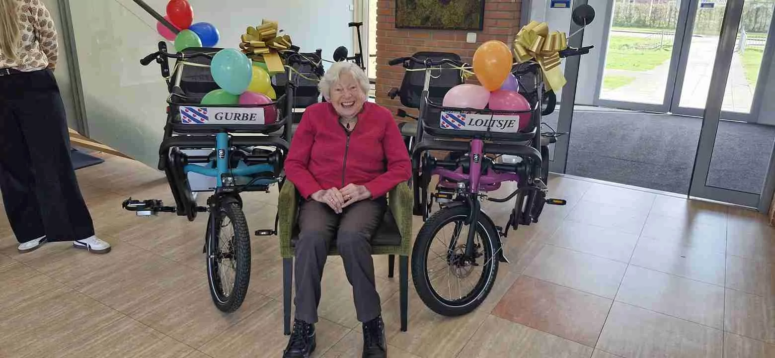 Een oudere vrouw zit glimlachend voor twee versierde driewielers met ballonnen en gouden strikken binnen. Op de naambordjes van de driewielers staat GURBE en Lotlyse. Twee mensen staan gedeeltelijk buiten beeld.