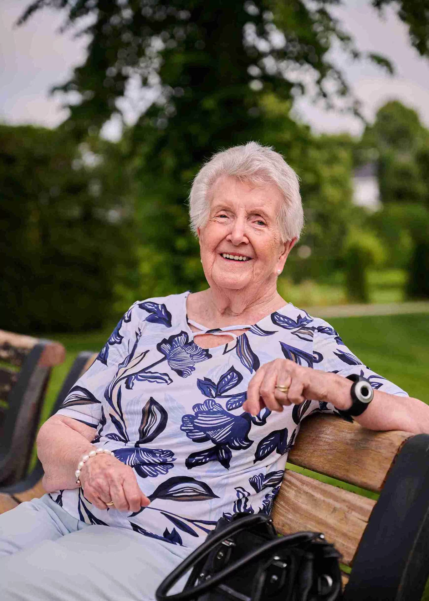 Een oudere vrouw met kort grijs haar zit glimlachend op een houten bankje in het park. Ze draagt een wit shirt met blauwe bloemmotieven, een lichte broek, een horloge en een zwarte handtas. Weelderig groen is zichtbaar op de achtergrond.