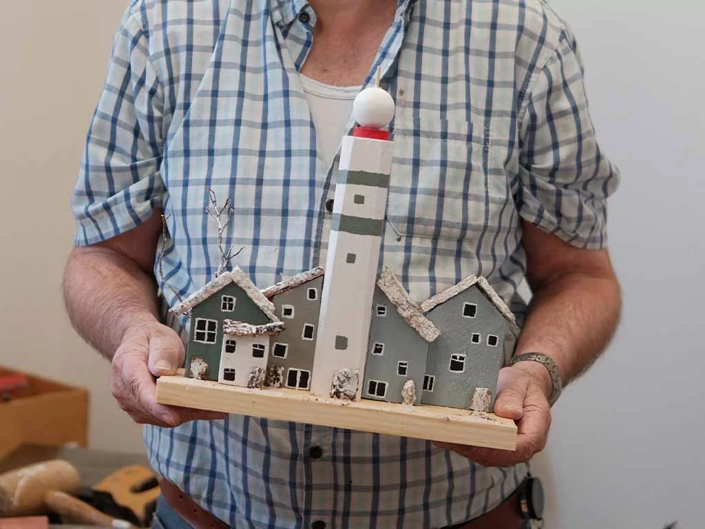 Een persoon in een geruit overhemd houdt een zelfgemaakt model van een vuurtoren vast, met aan weerszijden twee huisjes, allemaal op een houten voetstuk.