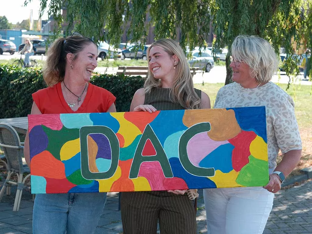 Drie vrouwen staan buiten, glimlachend, met een kleurrijk beschilderd bord in hun handen waarop in het midden in grote letters ‘DAC’ staat. Op de achtergrond zijn bomen en terrasstoelen te zien.