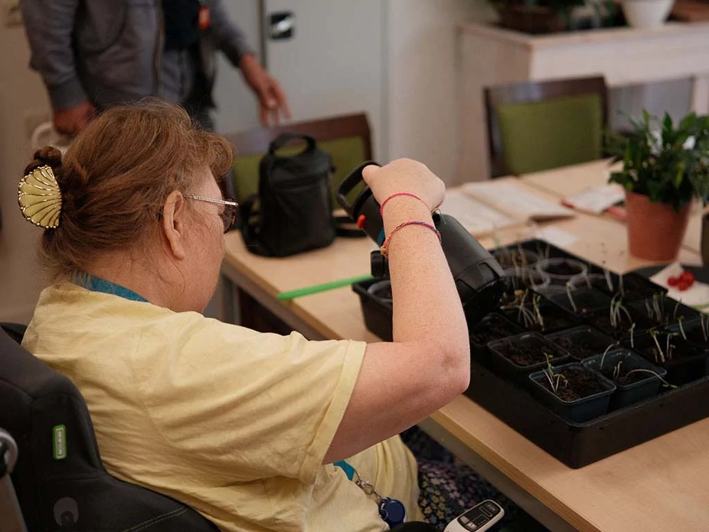 Een vrouw in een geel shirt geeft zaailingen in kleine potjes op een tafel water en verzorgt de planten binnenshuis.