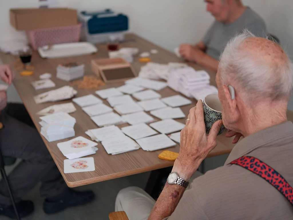 Drie ouderen zitten rond een tafel en sorteren papieren en enveloppen. Een man op de voorgrond houdt een mok vast. Op de tafel liggen netjes gerangschikte stapels papier en wat hapjes.