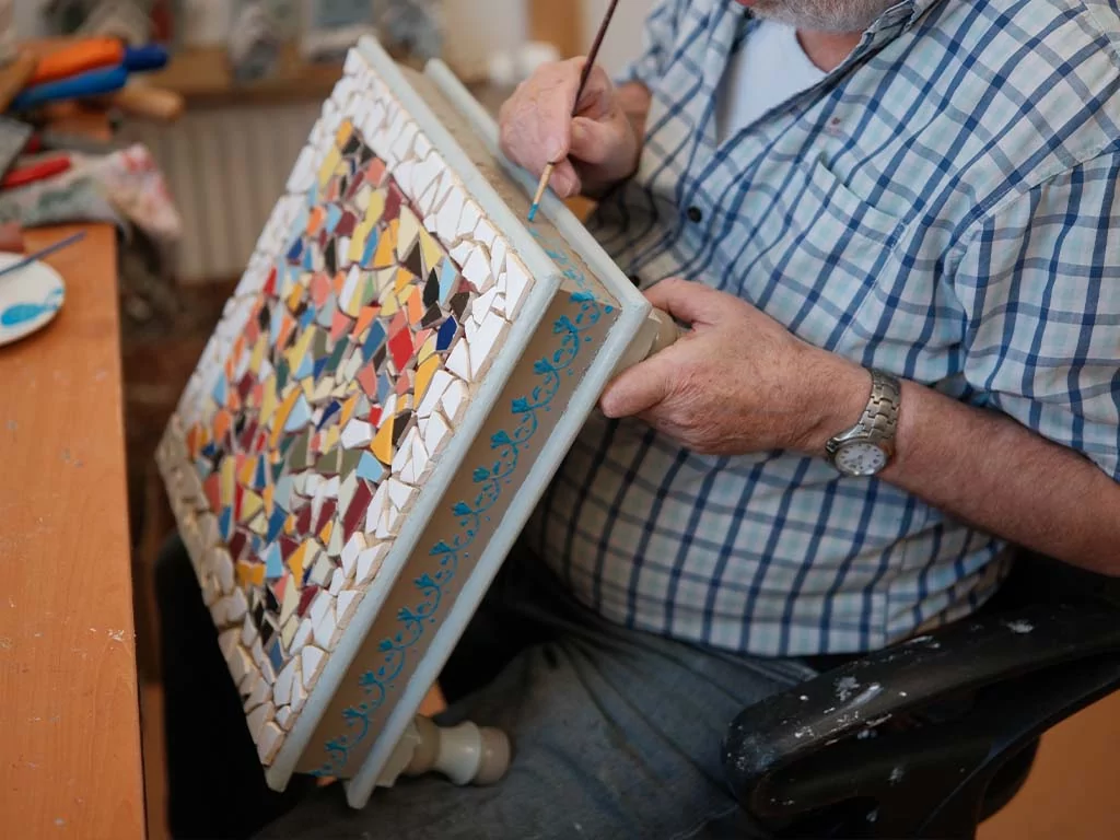 Een oudere man in een geruit overhemd schildert een blauw patroon op de zijkant van een rechthoekig mozaïekkunstwerk van kleurrijke gebroken tegels, terwijl hij aan een houten bureau zit.